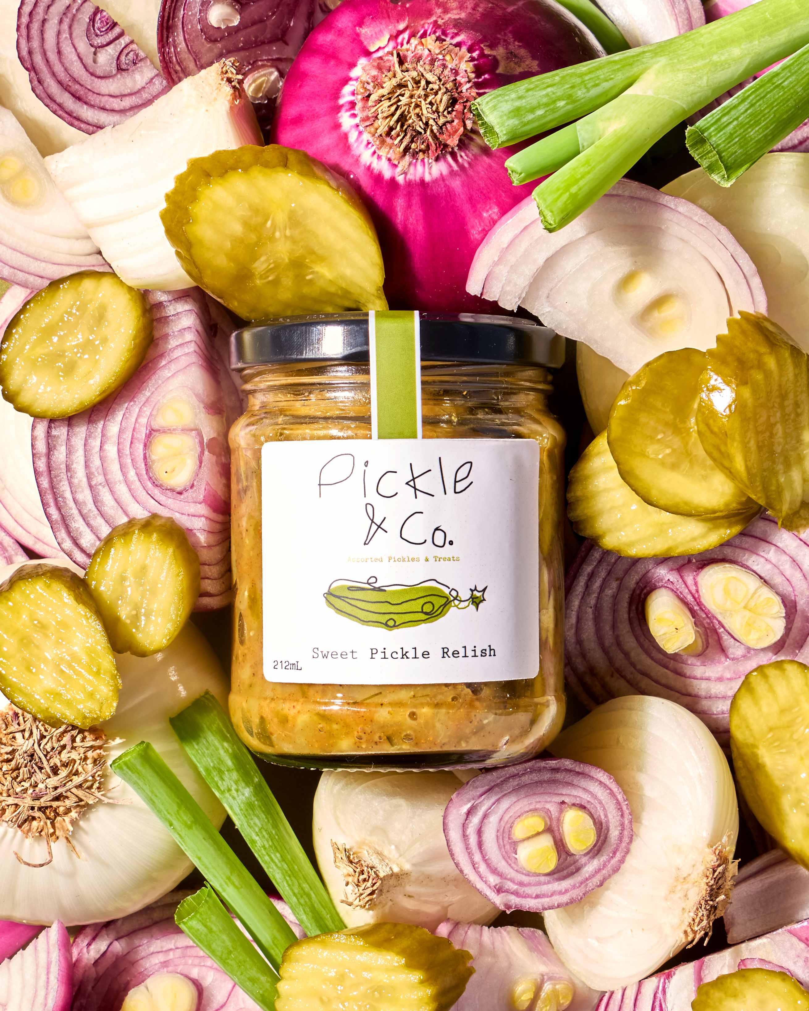 A jar of Pickle & Co. Sweet Pickle Relish nestled in sliced red onions, shallots and pickle chips